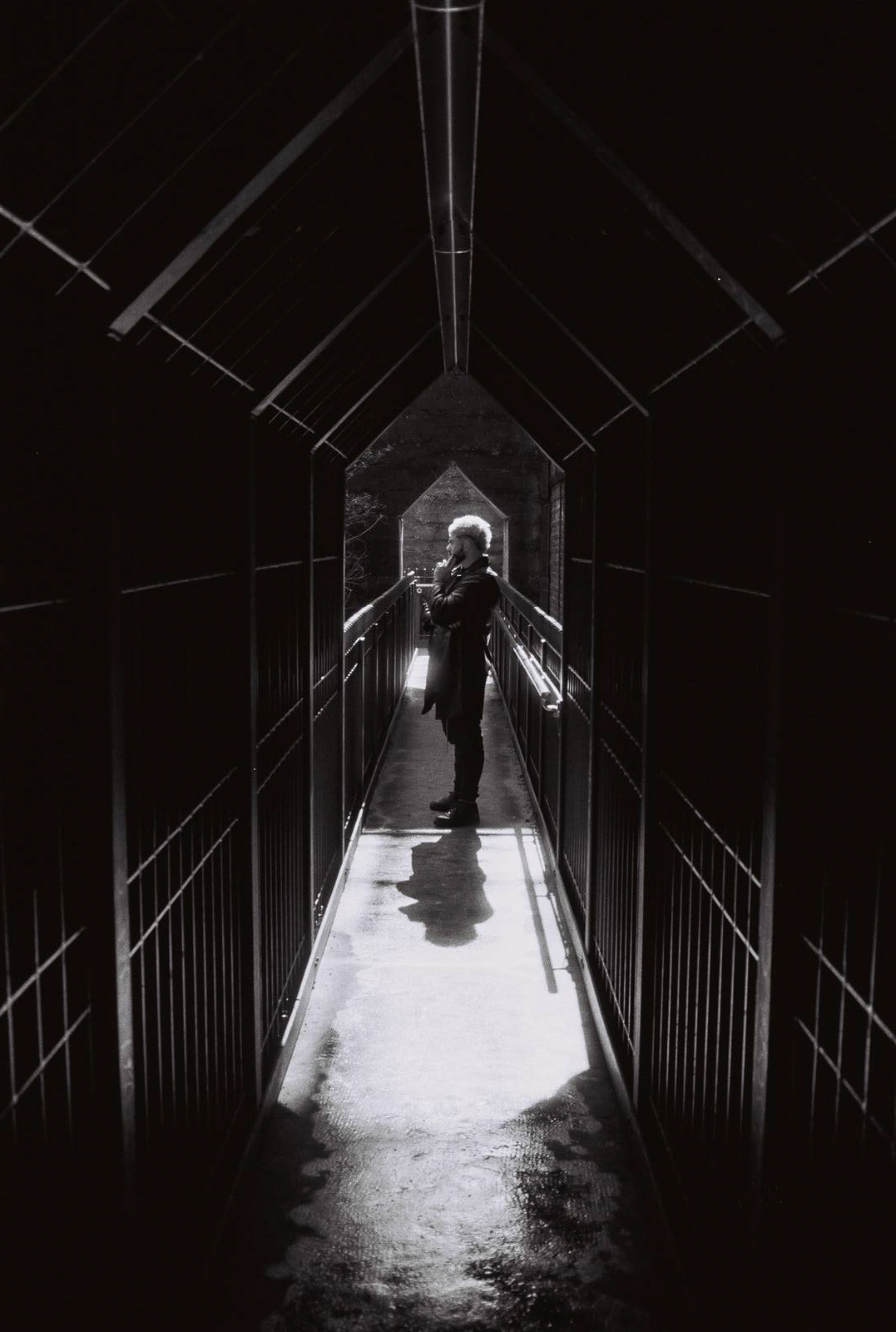A person silhouetted, standing in a dark, narrow corridor with metal railings, lit by a light source at the end.