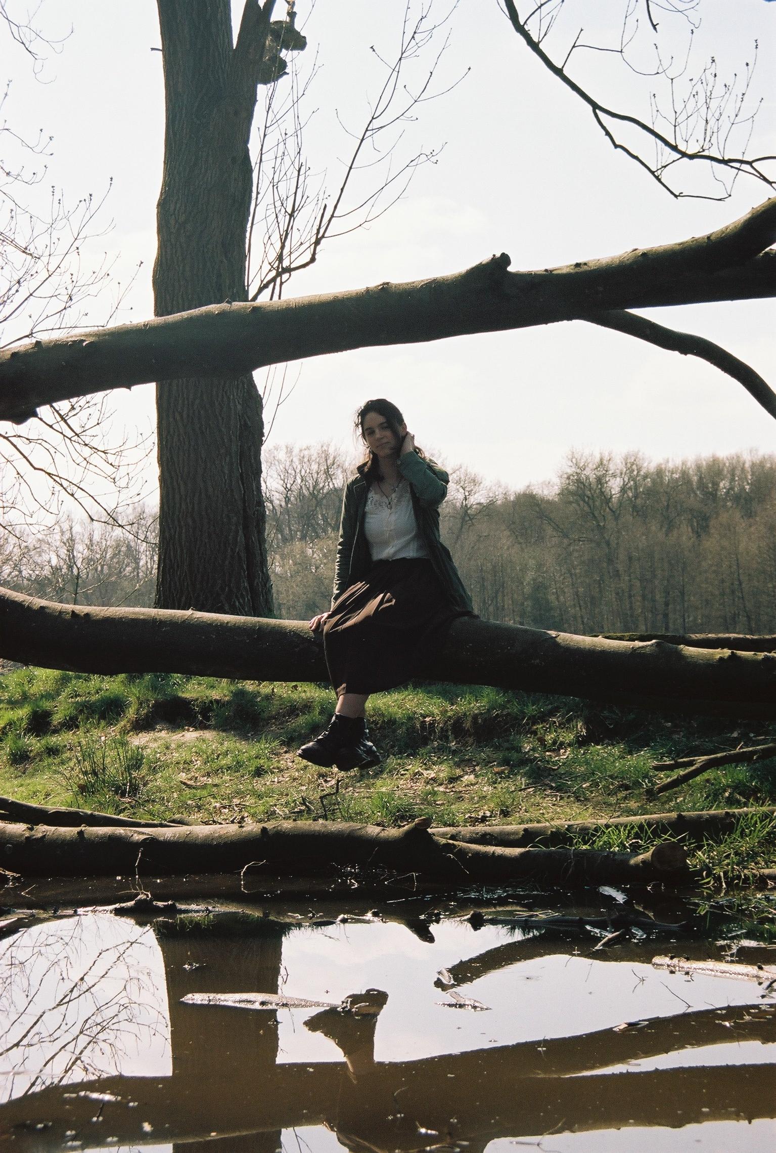 A young woman sits thoughtfully on a fallen tree branch over murky water in a wooded area. By NinoTurbaProductions