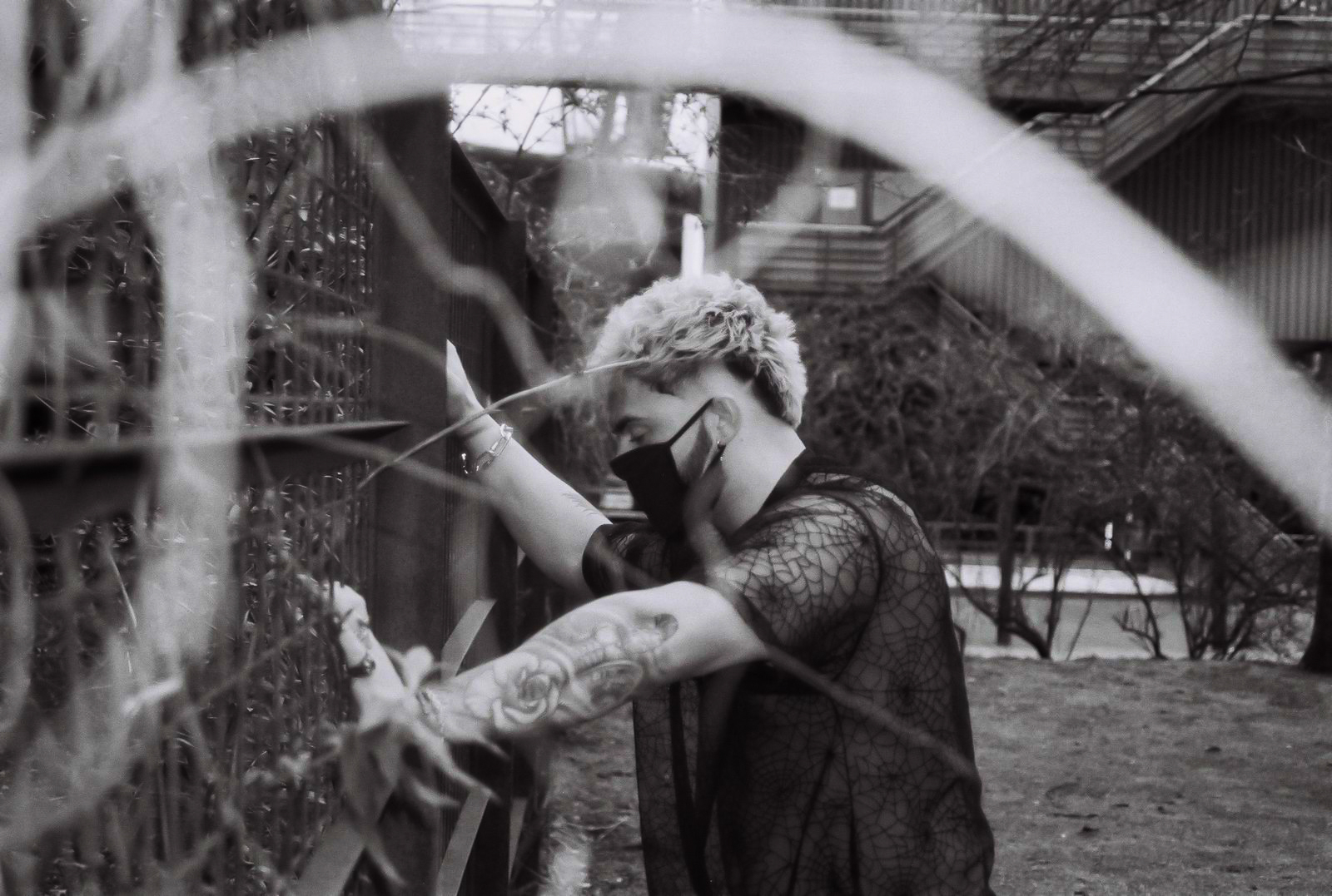 Portrait of a man leaning against a fence, with black, alternative clothing in an industrial setting. By NinoTurbaProductions