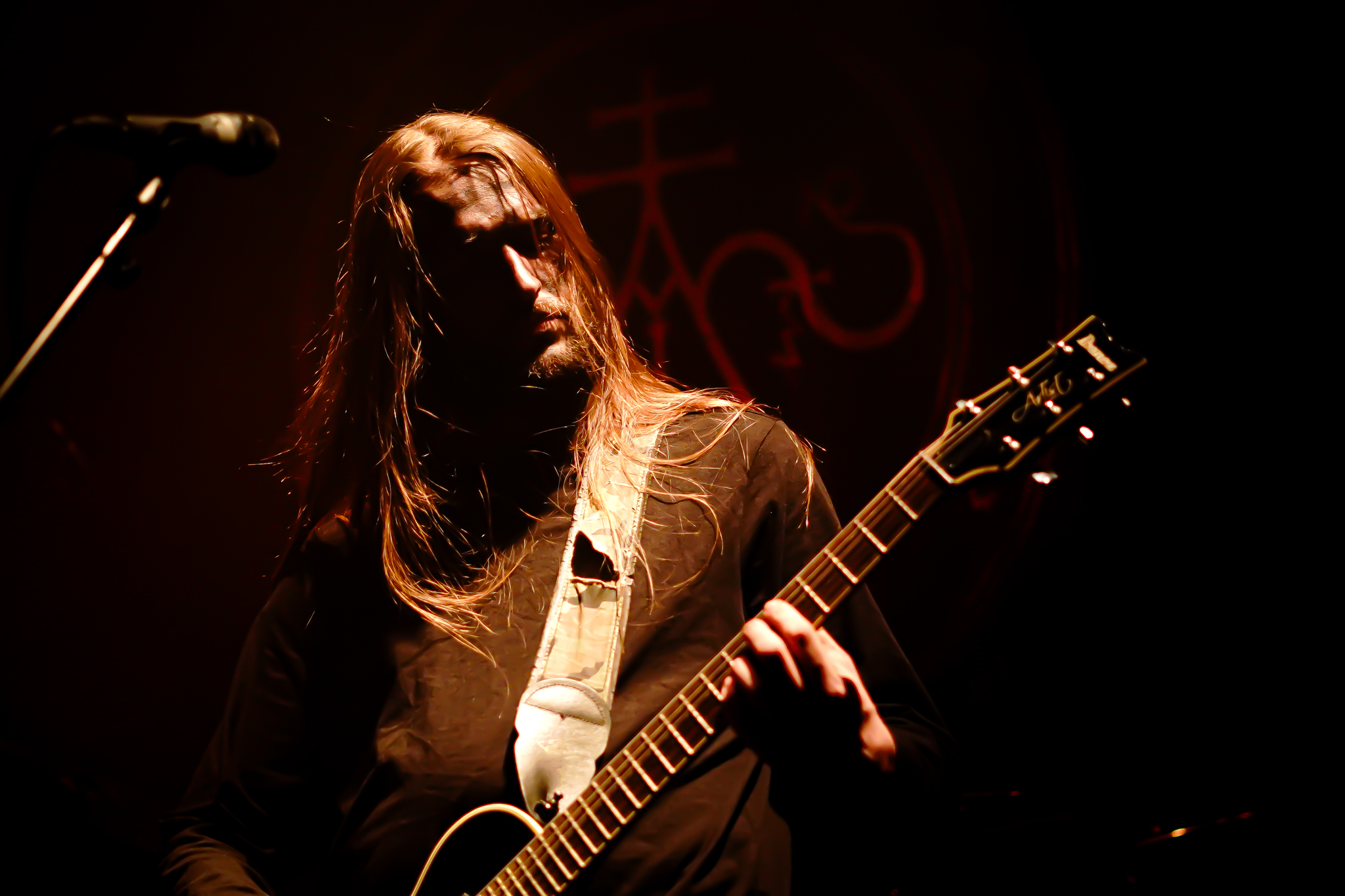 A person with long, dark hair, wearing a dark shirt, stands in a dark, moody setting.