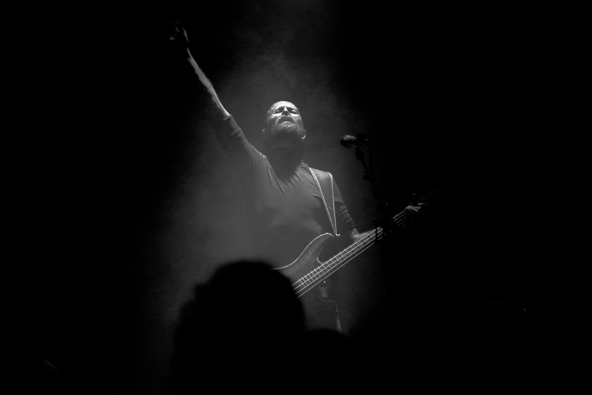 Musician lit by spotlight raises arm while playing bass guitar on stage, surrounded by darkness and faint smoke. Band: Shagor