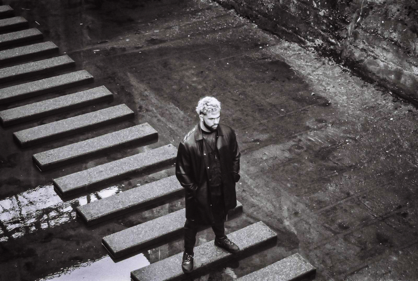 A man with white hair stands on concrete steps in a wet, dark, industrial-looking setting. By NinoTurbaProductions