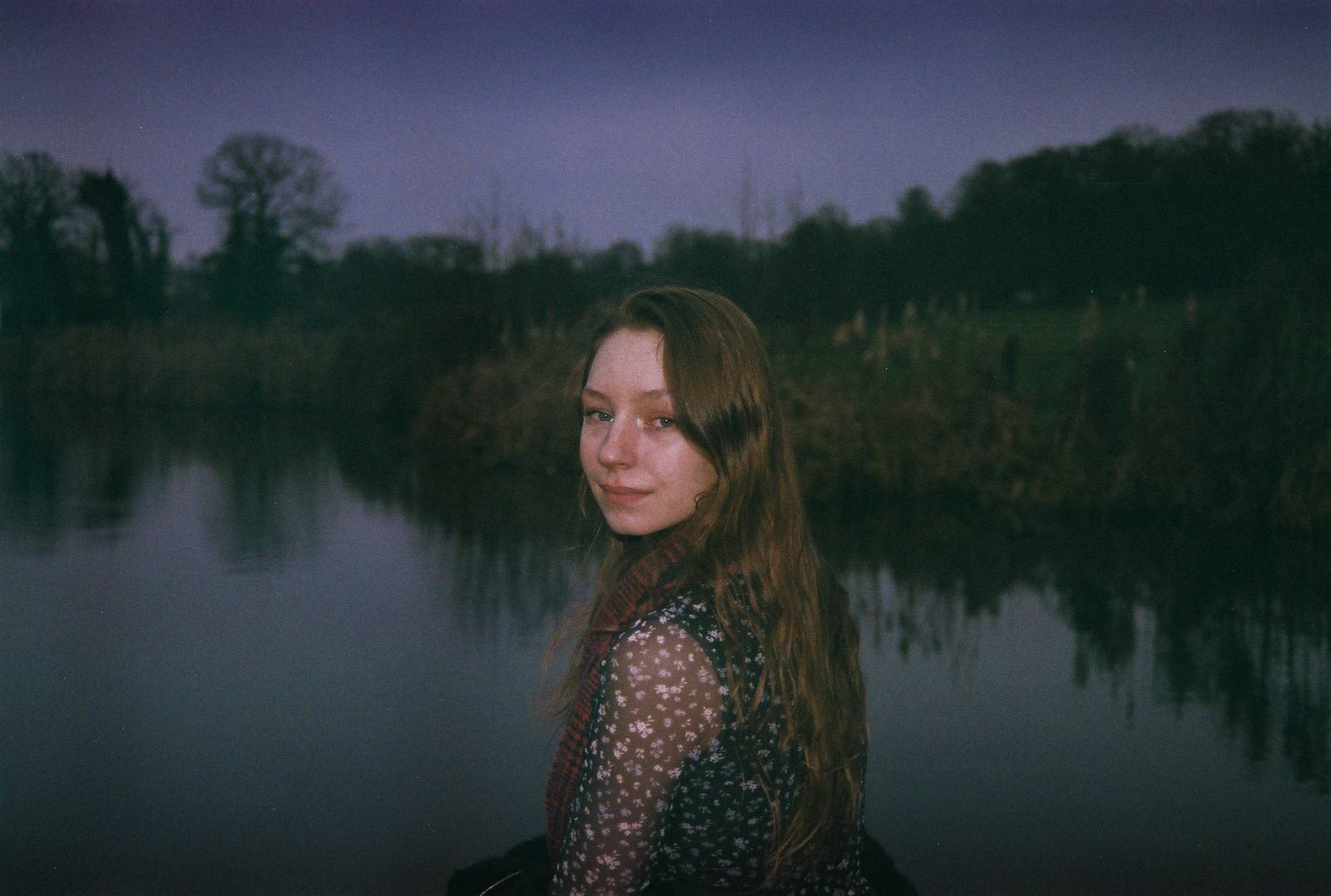 A young woman with long, brown hair smiles at the camera, with a dark, calm body of water behind her and trees.