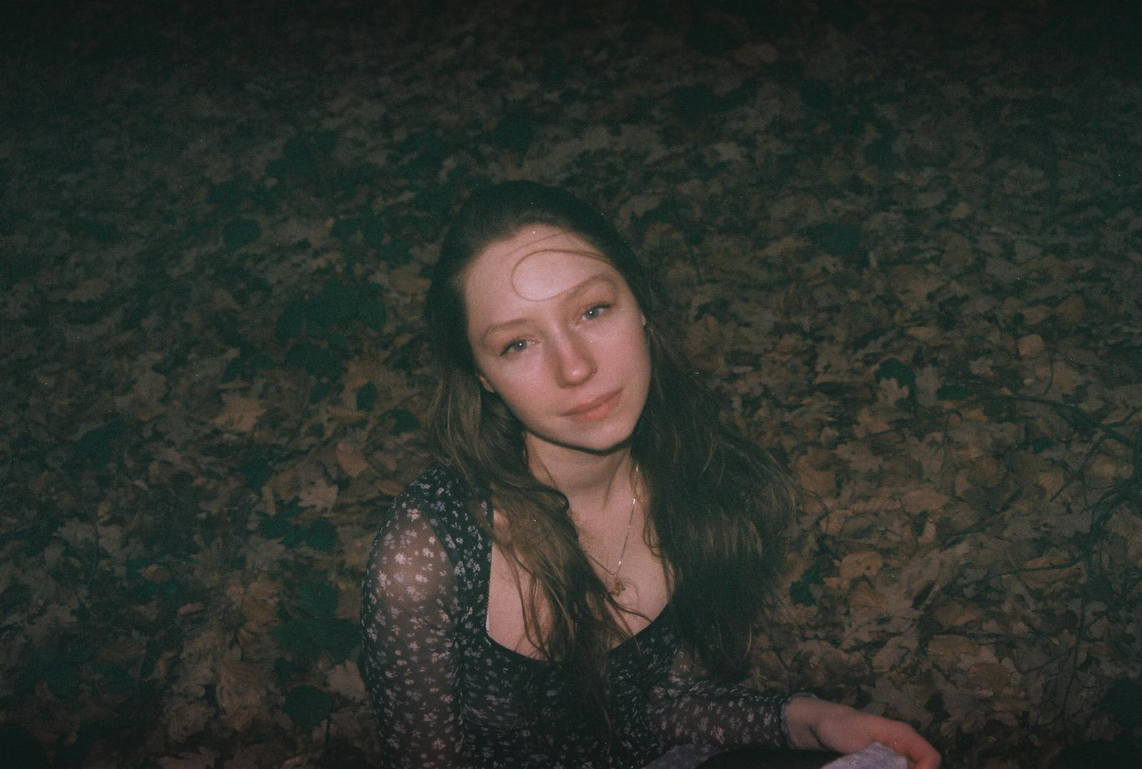 Young woman with long, dark hair in a floral top, sits among fallen leaves, looking softly at the camera.