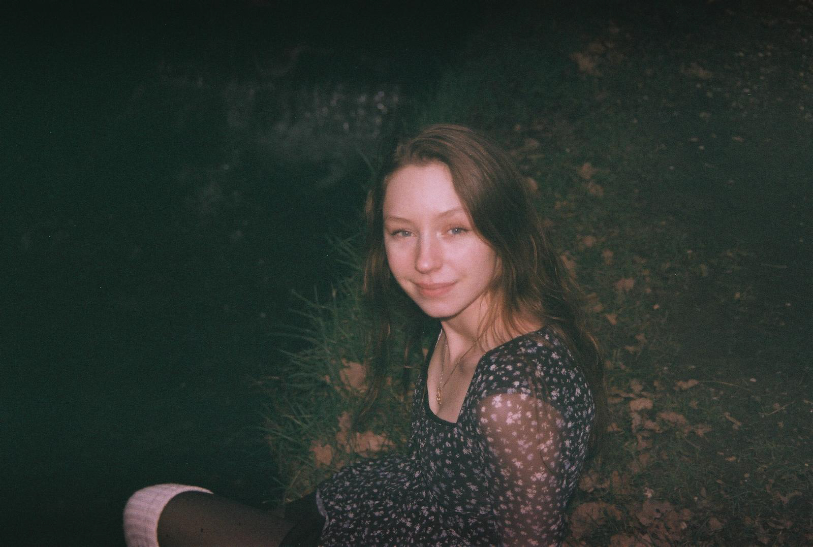 A young woman sitting next to a waterfall, among fallen leaves, smiles at the camera. By NinoTurbaProductions