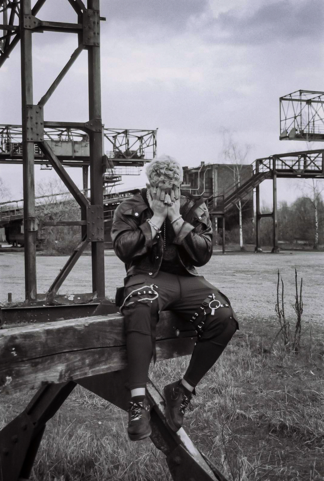 A man with light hair covers their face, sitting on a wooden beam in an industrial, outdoor setting. By NinoTurbaProductions