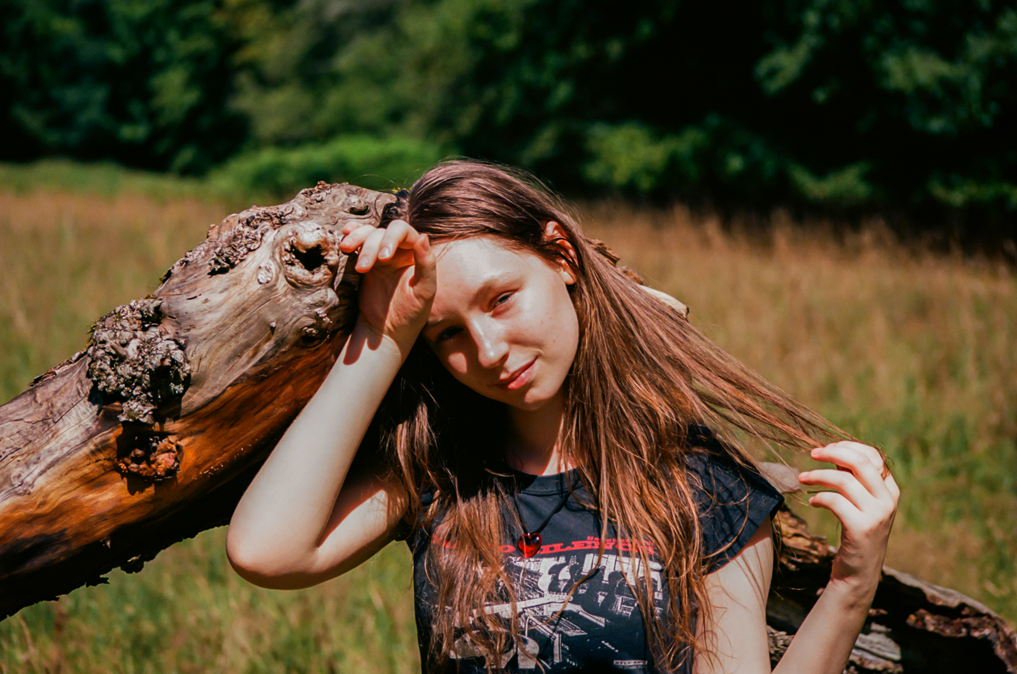 A young woman with long, brown hair leans against a large tree trunk, looking at the camera. By NinoTurbaProductions