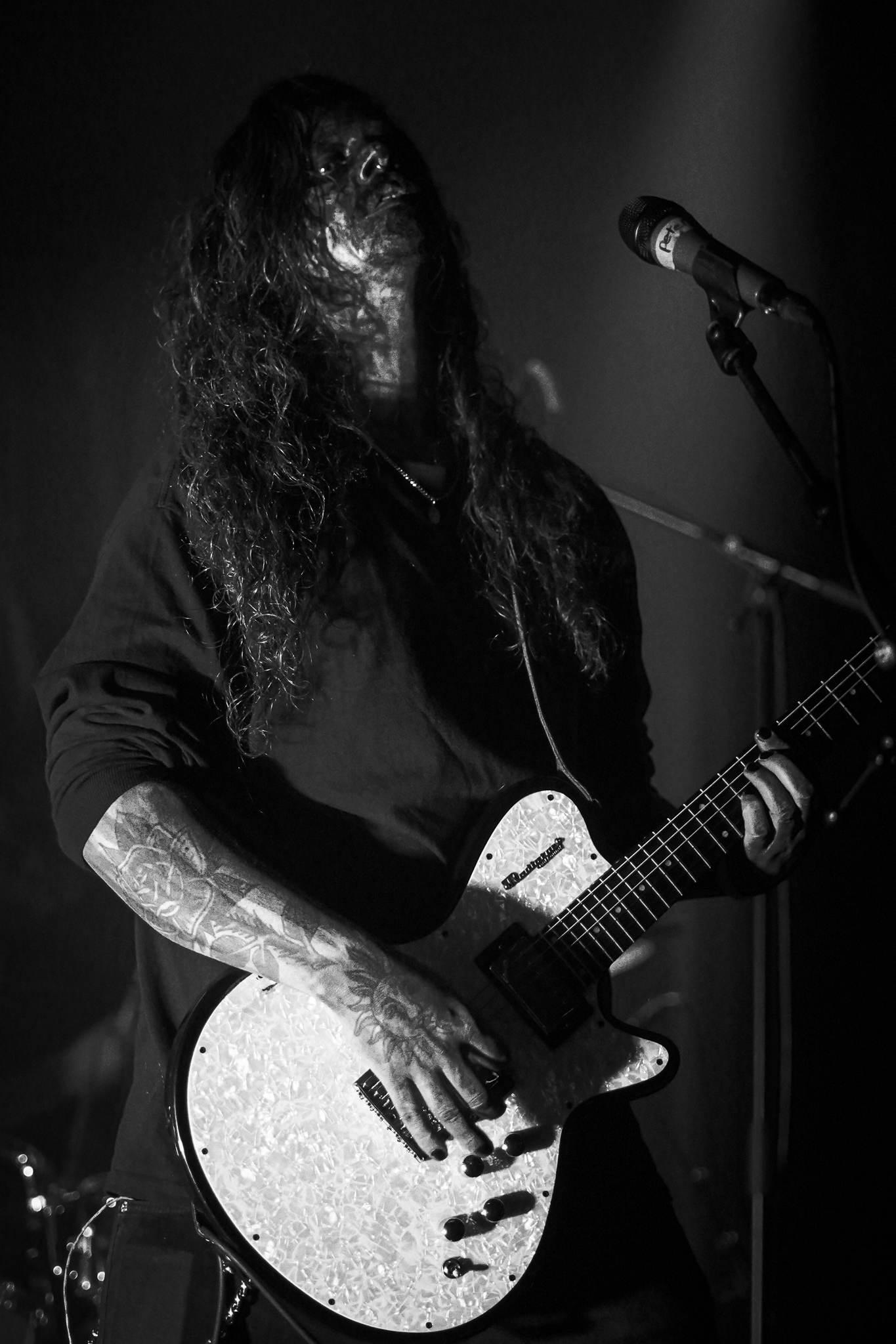 Photo of a long-haired guitarist with tattoos, performing under dramatic lighting, holding a guitar in front of a microphone