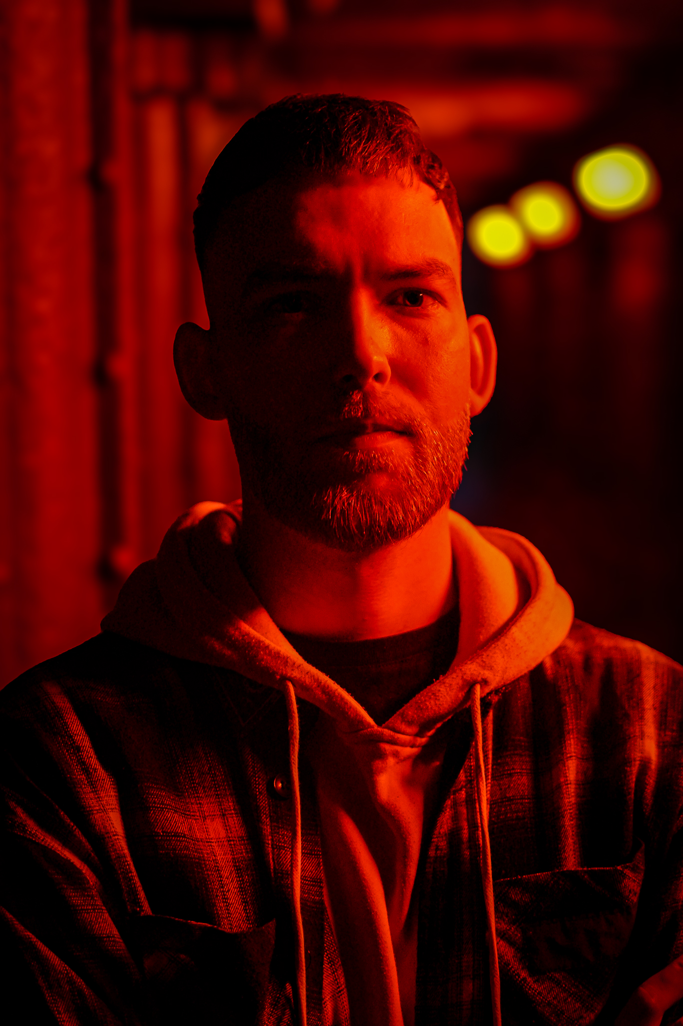 A portrait of a bearded man in a red light, wearing a hoodie and plaid shirt. By NinoTurbaProductions