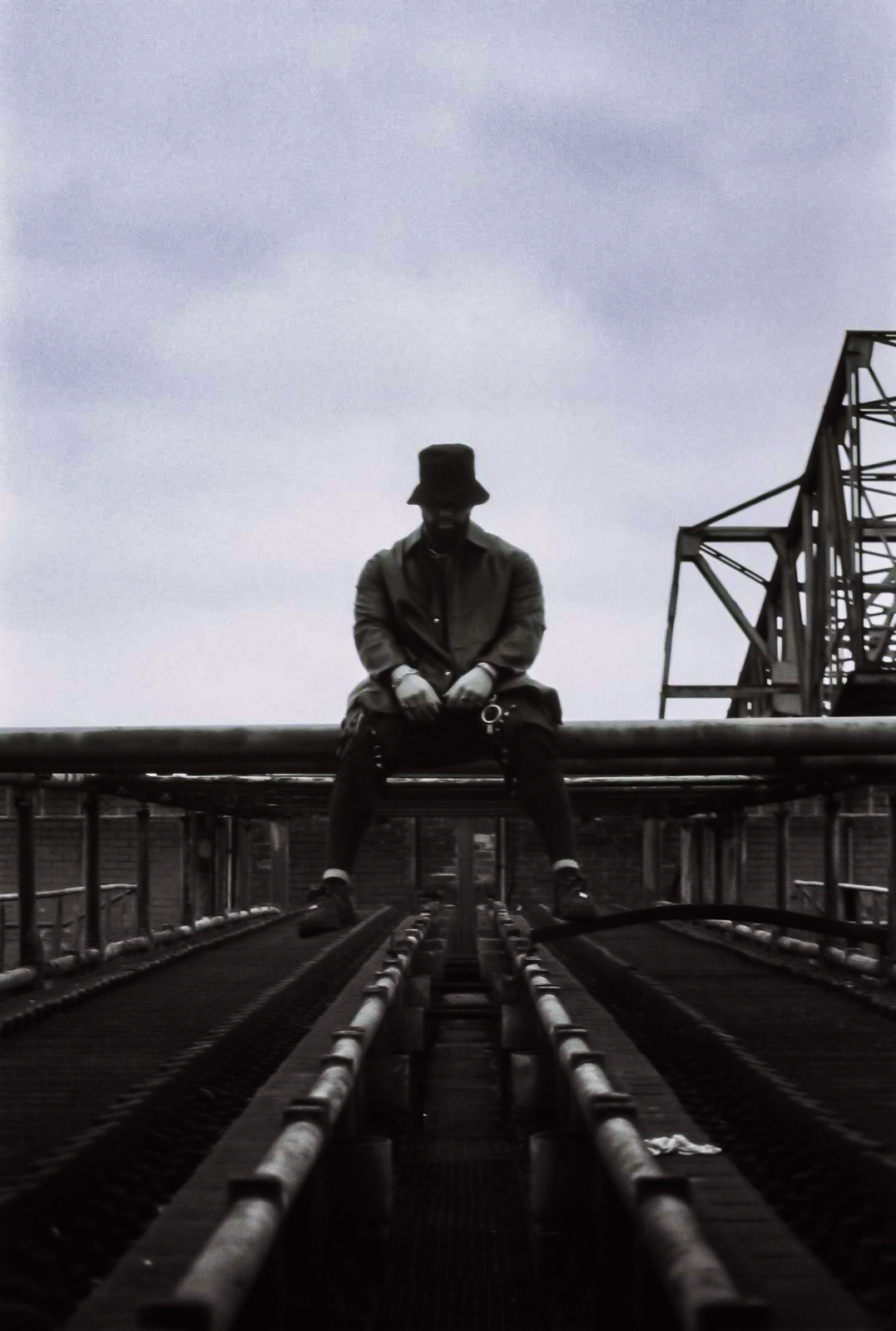 A man in a hat and coat sits on a metal structure, looking directly ahead, under a cloudy sky. By NinoTurbaProductions