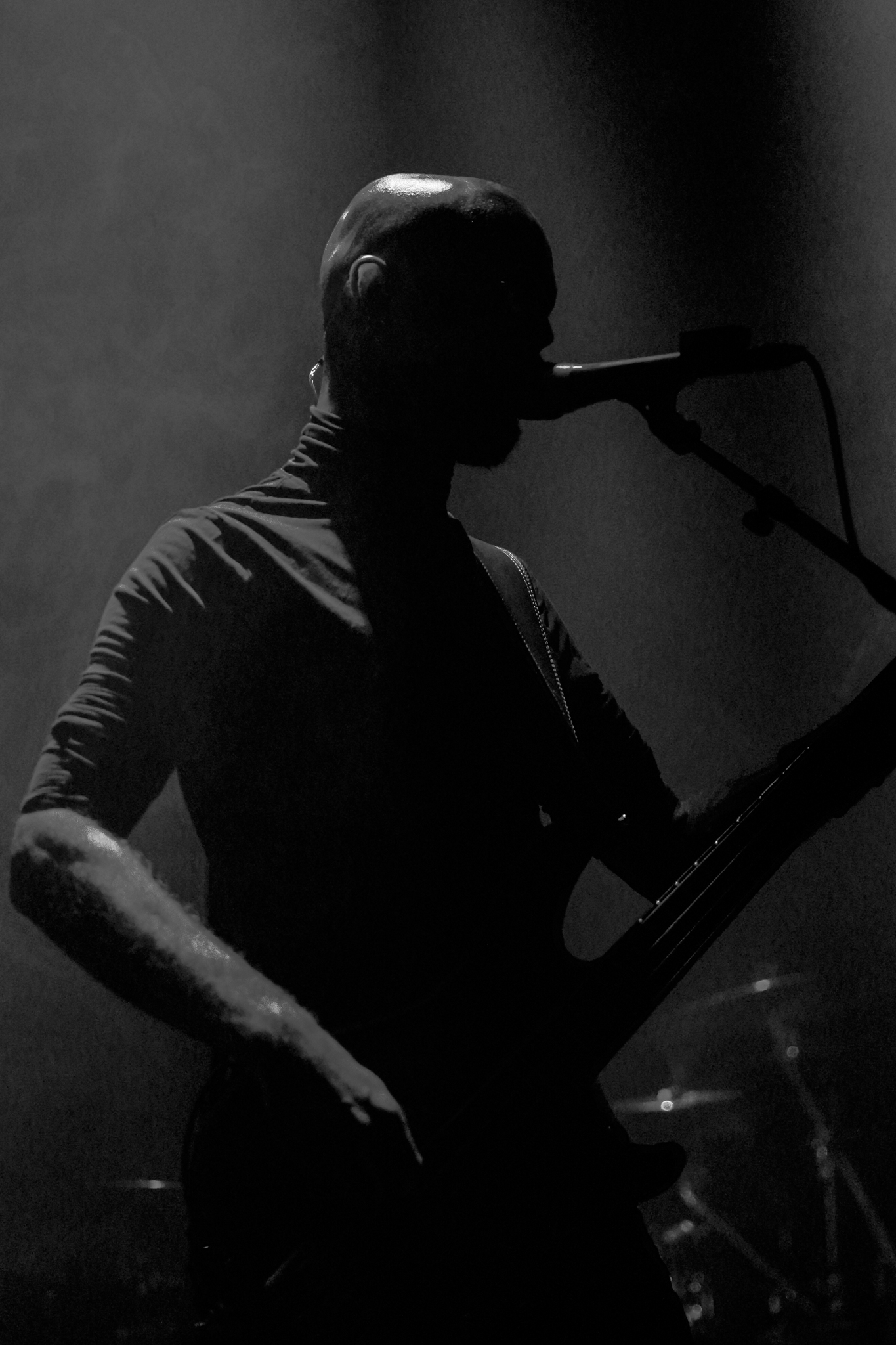Silhouetted bassist with a bald head performing on a dimly lit stage, singing into a microphone with drums in the background.
