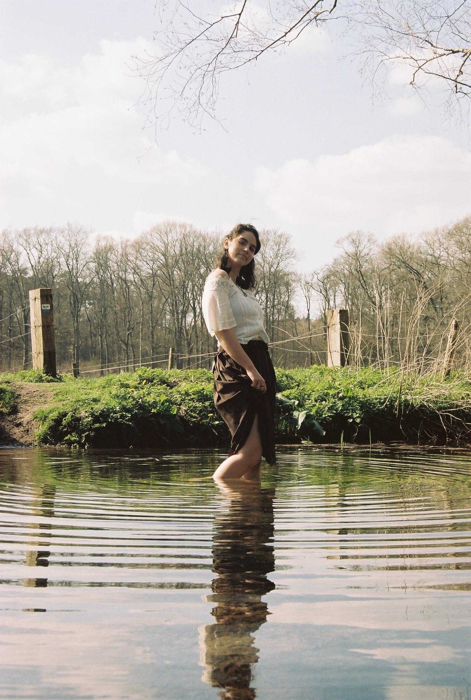 A young woman stands in shallow water, smiling at the camera, with a forest in the background. By NinoTurbaProductions