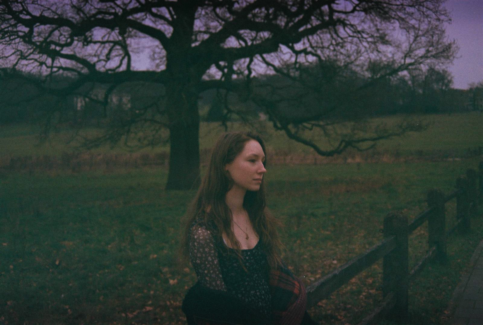 A young woman, lost in thought in a park. A big tree is visible behind her. By NinoTurbaProductions