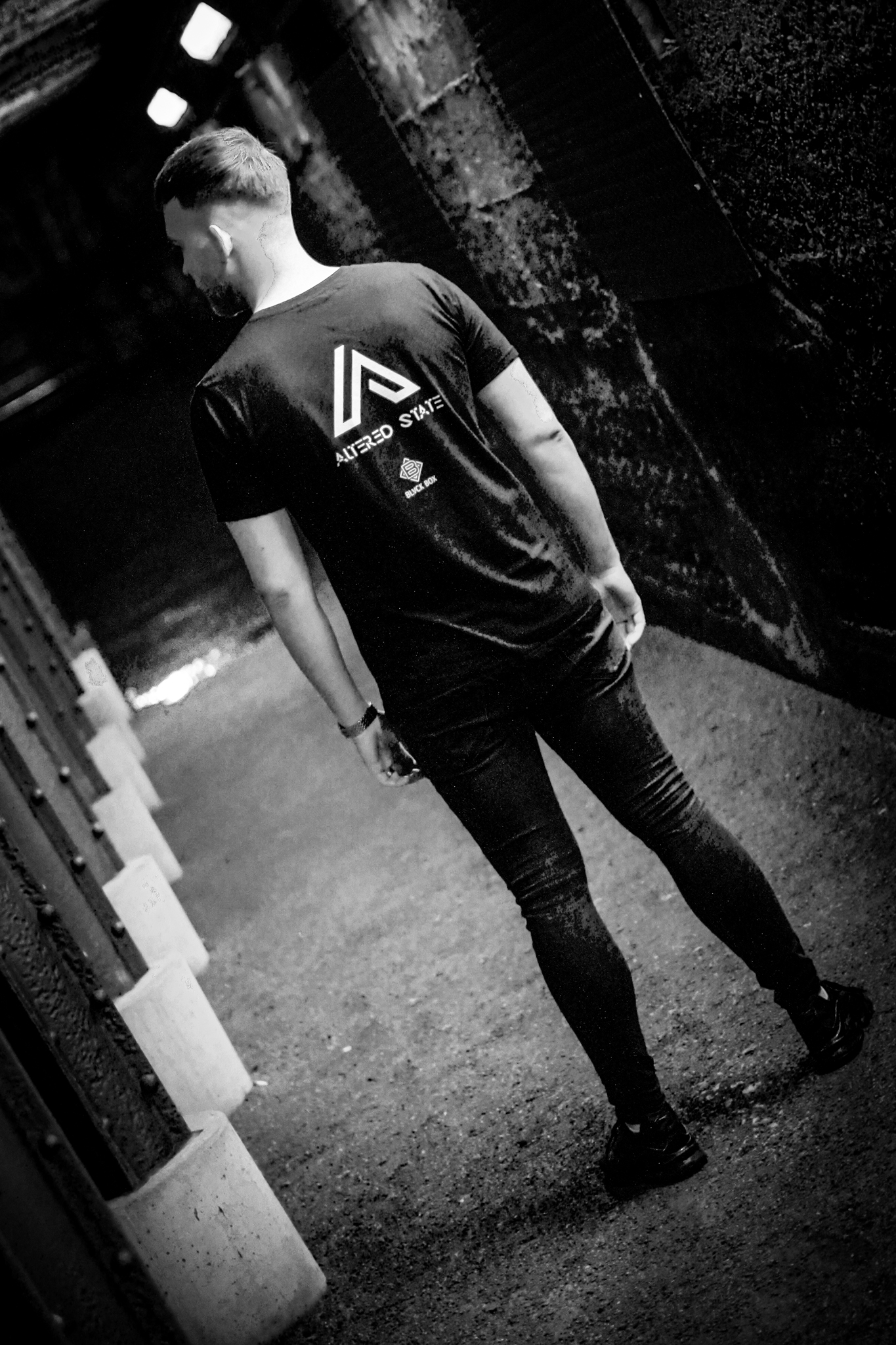 A man in a black t-shirt with ALTERED STATE on it, looks down a narrow, metal tunnel. By NinoTurbaProductions